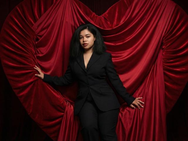 SunshineFlow in a black suit, standing with arms against red, heart-shaped, satin curtains. She has black hair, red lipstick, and a confident expression. Her pussy is not visible.
