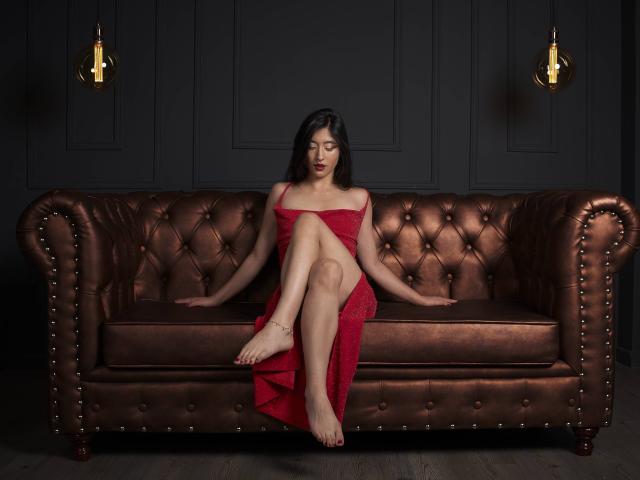 TaharaRomanov sits on a brown leather couch, wearing a red lace dress, exposing her tits and pussy, legs crossed, arms resting on the couch, bare feet on the floor.