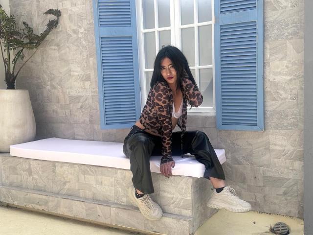 TaharaRomanov with long black hair, leopard print cardigan, black leather pants, and white sneakers, sitting on a white cushioned bench, blue shutters, grey stone wall, potted plant.