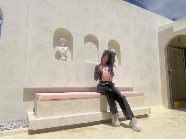 TaharaRomanov with black hair, wearing a leopard print top, exposing small boobs, black pants, and white boots, sitting on a pink bench against a white wall with statues.