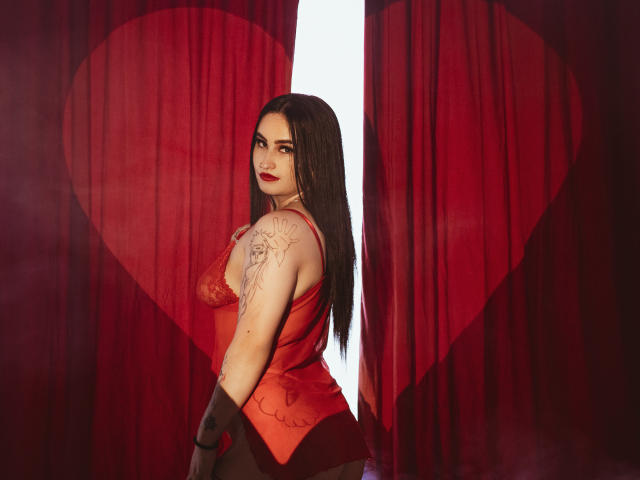 ScarletPriynce with long black hair, red lace top, tattoo on right arm, black gloves, looking over shoulder, red curtains with heart, red and black colors, sexy, confident.