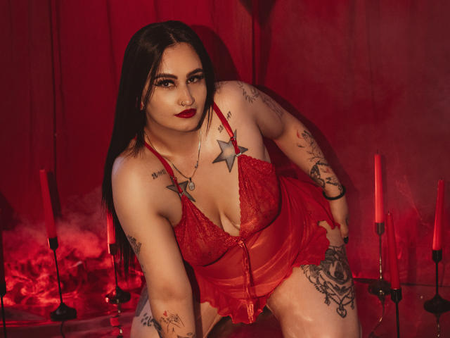 ScarletPriynce with long dark hair, red lace lingerie, tattoos, red lipstick, and a red star tattoo on her chest, leaning forward, hands on thighs, surrounded by red candles.
