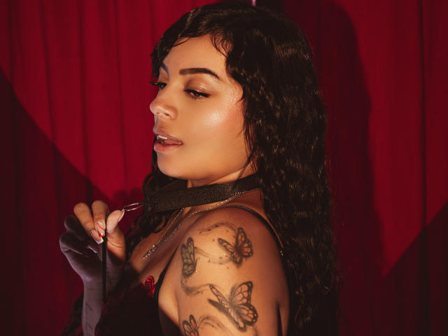 KateMayoss with curly black hair, holding a black strap, has butterfly tattoos on her shoulder, wearing a black top, and has a sexy, confident expression, red curtain background.