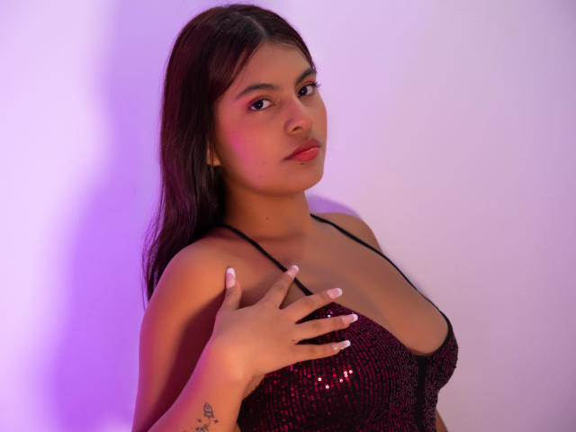 AlyssaGreen with long brown hair, pink eye makeup, and a black sequined top, touching her tits with a hand, has a tattoo on her right wrist, and a purple glow.