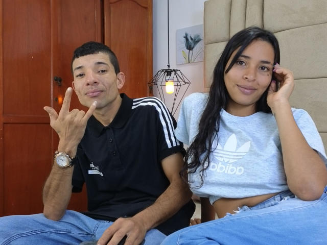 JazminaAndJosephe, sitting together, she with long black hair in a gray Adidas shirt, he with short black hair in a black Adidas shirt, making a "rock on" hand sign.