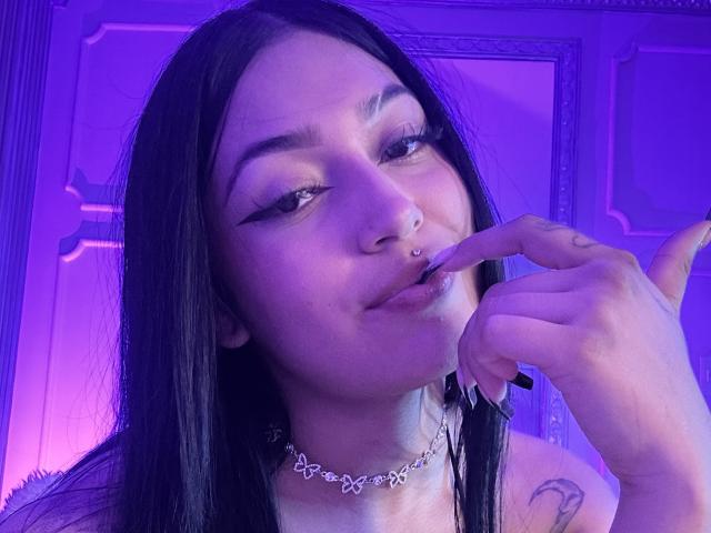 KatianPierce with black hair, nose ring, and butterfly necklace, biting her finger, showing off long black eyeliner and a tattoo on her left shoulder, under purple lighting.
