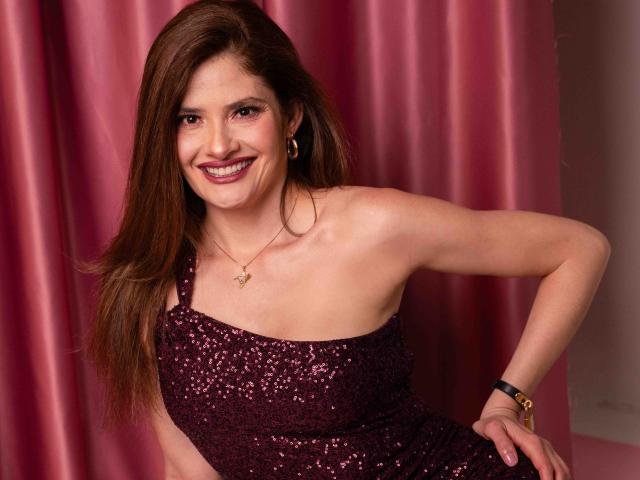 CelineBoyer with long brown hair, smiling, wearing a sparkly purple one-shoulder dress, showing her bare shoulder and a gold necklace, standing against pink curtains.