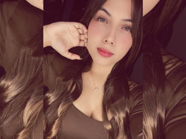 NailaIsaa with long, wavy brown hair, pink lips, and a brown top, showing off her tits with one hand on her head, wearing a delicate necklace.