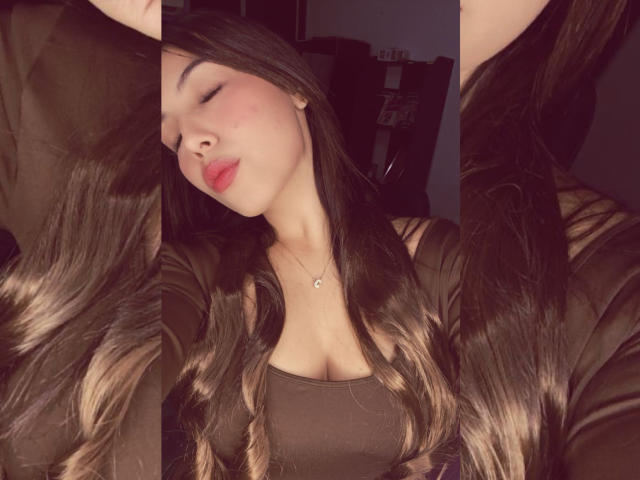 NailaIsaa with long, dark brown hair, slightly wavy, wearing a black top, eyes closed, red lipstick, small necklace, soft smile, in a dimly lit room, arms raised.