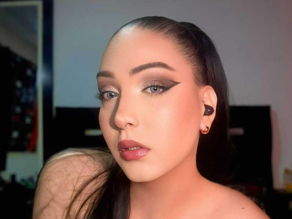 BlessExotic has shiny, light brown skin, thick black eyebrows, winged eyeliner, and glossy pink lips. She has long black hair in a ponytail, and black ear gauges.