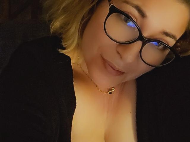 MimiCoquinne with glasses, blonde wavy hair, black top, cleavage, subtle smile, gold necklace, soft light, cozy atmosphere.