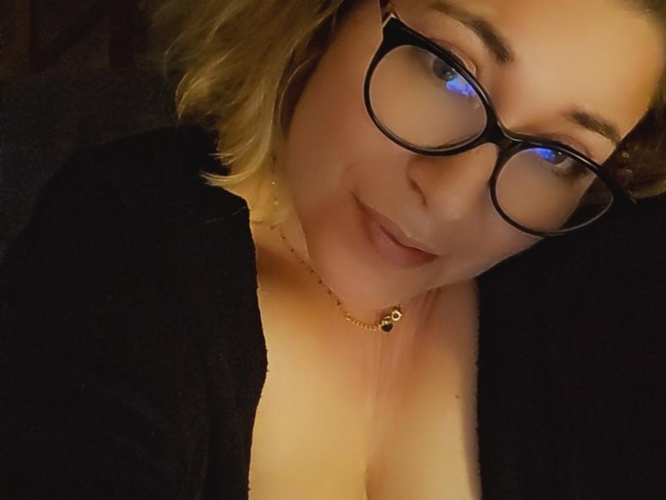 MimiCoquinne with glasses, blonde wavy hair, black top, cleavage, subtle smile, gold necklace, soft light, cozy atmosphere.