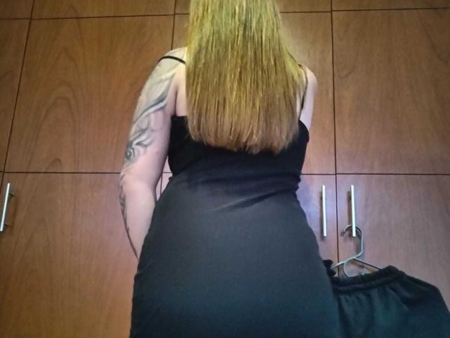 Catag has long blonde hair, black dress, tattoo on left arm, and a curvy ass, standing with back to camera, wooden cabinet in background, black bag on right.