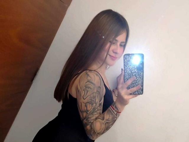 Catag with long brown hair, black dress, tattooed arm, holding phone, taking mirror selfie, showing ass, small breasts, smiling, phone flash, white wall, wooden door.