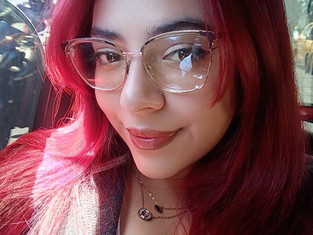 Close-up of RousseColliinss with bright red hair, wearing clear, rectangular glasses and glossy red lipstick. Sunlight reflects in her glasses, highlighting her dark eyebrows and smooth skin. She has a subtle, confident smile. A gold necklace with a small, circular pendant rests on her chest. The texture of her hair is smooth and slightly wavy. The image has a natural, vibrant feel.