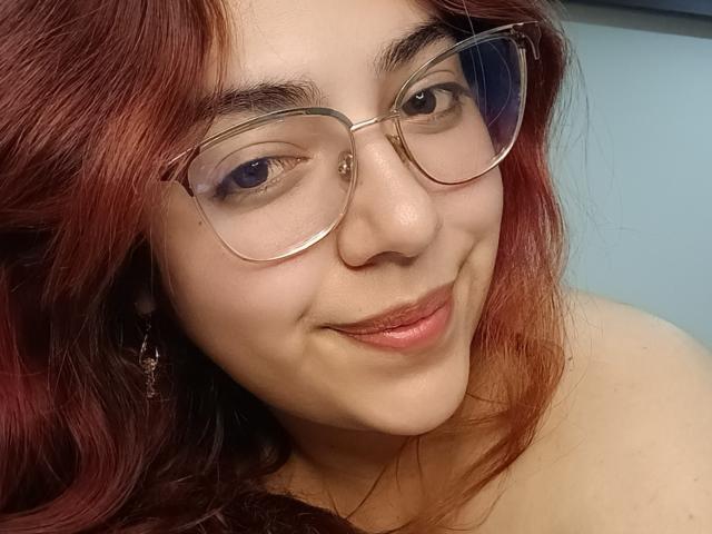 RousseColliinss with red hair, glasses, and a slight smile, showing her cute nose and glossy pink lips. Her tits are partially visible, and she's wearing a dangle earring.
