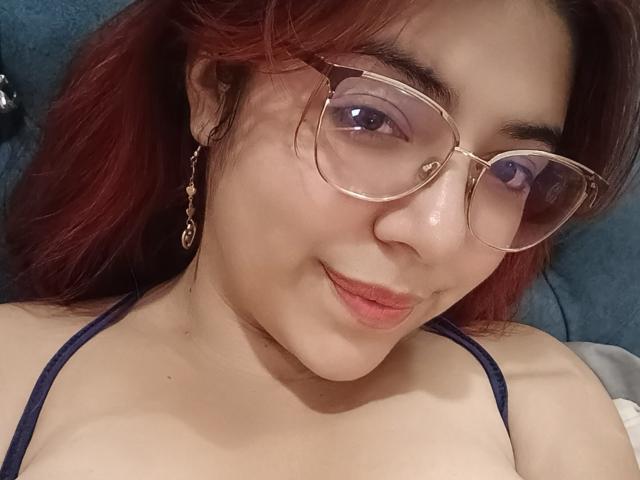RousseColliinss with red hair, wearing glasses and a dark strap top, showing her tits and a slight smile, lying down with a cute, relaxed expression.