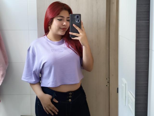 RousseColliinss with red hair, wearing a lilac crop top and black high-waisted jeans, holding a phone in a mirror selfie, showing her tits and a hand on her ass.
