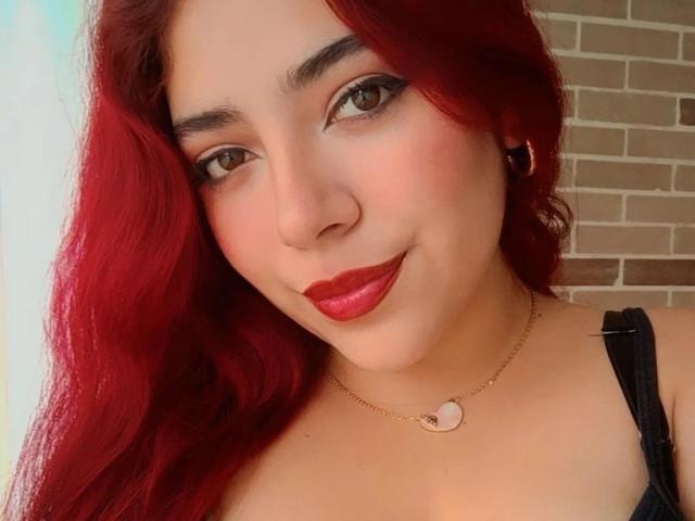 RousseColliinss has vibrant red hair flowing to the left, with light brown eyes and subtle black eyeliner. She wears bold red lipstick and a small gold necklace with a heart-shaped pendant. She has a light skin tone and small gold earrings. Her black strap is visible on her left shoulder. The image focuses on her face, which is slightly tilted to the right. The texture of her hair is smooth, and her expression is a gentle smile.