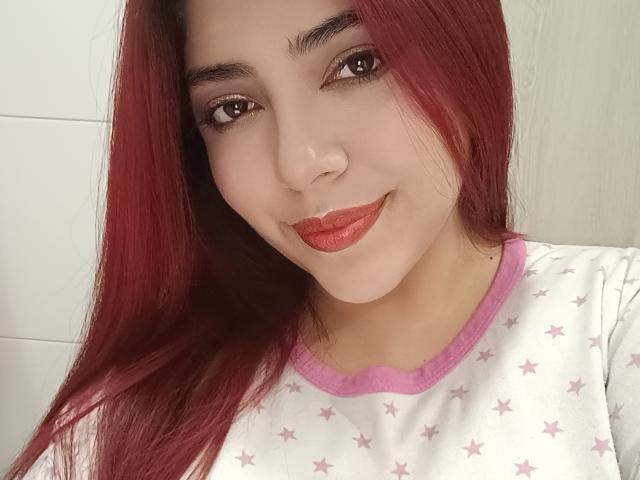RousseColliinss with red lipstick, straight red hair, and pink-star pajama top, smiling at the camera, showing her cute face and small ass. Her brown eyes are slightly upturned.