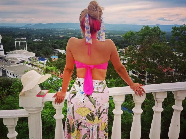 AdamsMerlina's back is to the camera, wearing a pink halter top and floral skirt, standing on a balcony with hands on the railing, hat on the rail.