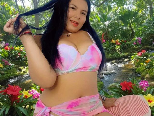 Olivia Carson, con cabello negro, un bikini de tie-dye rosa y blanco, sujetándose el cabello, sonriendo, mostrando sus grandes senos, una falda envolvente rosa, un fondo de bosque verde, flores coloridas y un pequeño arroyo..