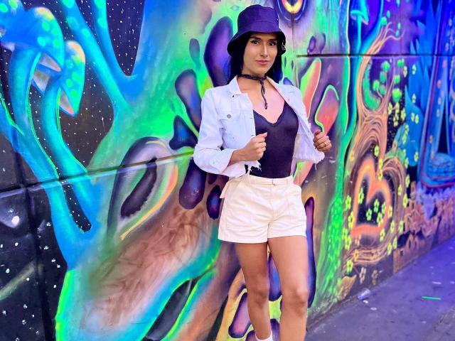 FallonRogers with tits in a black top, white shorts, and a blue bucket hat, standing against colorful graffiti, wearing a light blue jacket and black choker.