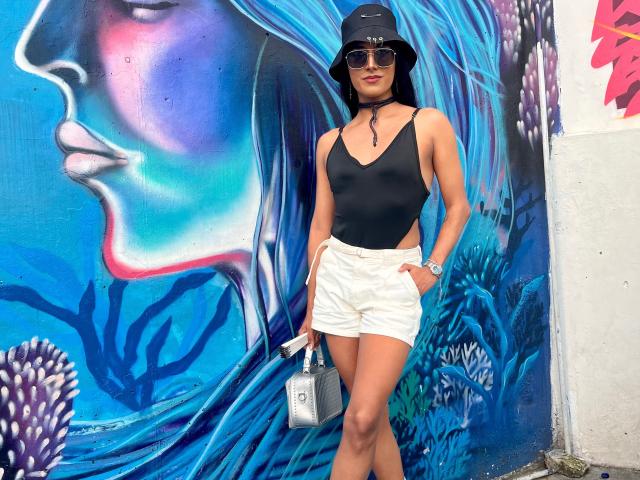 FallonRogers with taut ass in black top, white shorts, black bucket hat, sunglasses, holding silver bag, standing against blue graffiti of a woman's face.