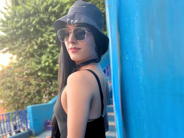 FallonRogers with long black hair, wearing a black cap, sunglasses, and a black tank top, showing her ass in a blue-walled outdoor setting with green trees.
