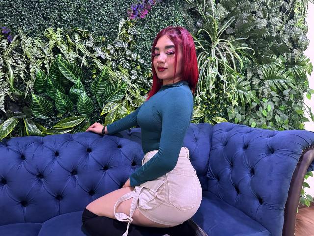 MiilaRouge with red hair, green long-sleeve top, and white high-waisted shorts, kneeling on a blue tufted couch, showing her big ass and small white garter straps.