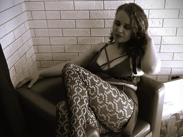A sepia-toned photograph of VittoriaI, a woman with curly dark hair, sitting in a leather armchair. She wears a black lace bra and patterned pants with white floral designs. Her left hand rests on the armchair's arm, while her right hand touches her hair. She looks down with a slight smile. The textured white brick wall is in the background. Light from the right side casts subtle shadows. The overall composition is relaxed and casual.