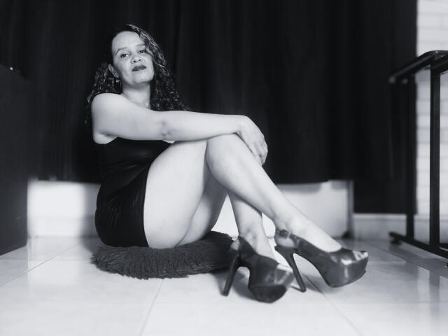VittoriaI with curly hair, sitting on a furry mat, wearing a black dress, and high heels, legs crossed, arms resting on knees, looking slightly up. Her ass is visible.