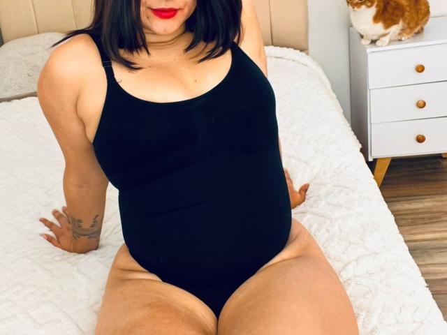 Carolly with black bob, red lipstick, black tank top, showing her tits and pussy, sitting on a white bed, tattoo on left arm, cat on right dresser.