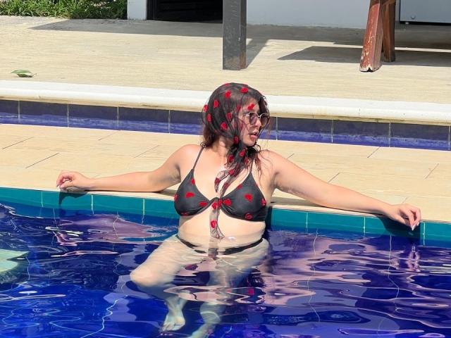 LinaFoxyHot in a blue-tiled pool, wearing a black bikini with red heart patterns, her tits visible, arms resting on the edge, water splashing on her face.