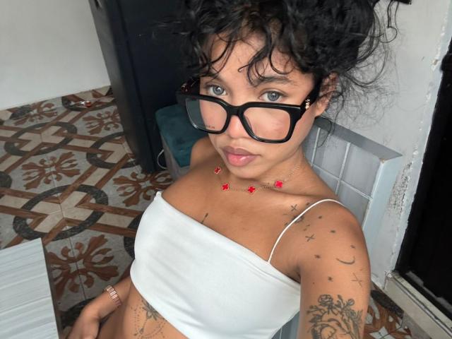 KathaThoms with curly black hair, black glasses, white tube top, and red bead necklace, sticking out tongue, tattoos on arm and chest, sitting indoors, showing small tits.