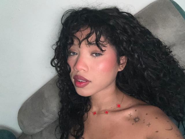 KathaThoms with curly black hair, pink blush, red lipstick, small red flower necklace, sun tattoo on right shoulder, lying on grey couch, looking at camera, topless.