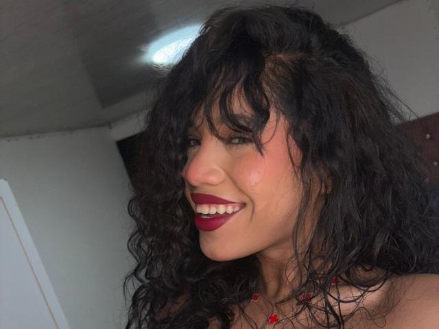 KathaThoms has curly black hair, red lipstick, and is smiling at the camera with visible teeth, wearing a red necklace. Her ass is not visible.