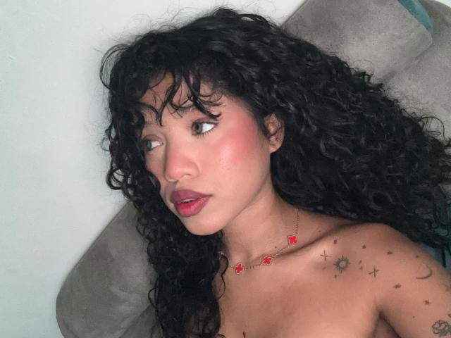 KathaThoms has curly black hair, pink blush, red lipstick, and small tattoos on her shoulder. She's wearing a red necklace with tiny flowers and looking to the side.
