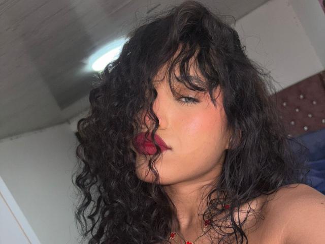 KathaThoms with curly black hair, red lipstick, and fair skin, looking at the camera, her tits barely visible, in a dimly lit room with a ceiling light.