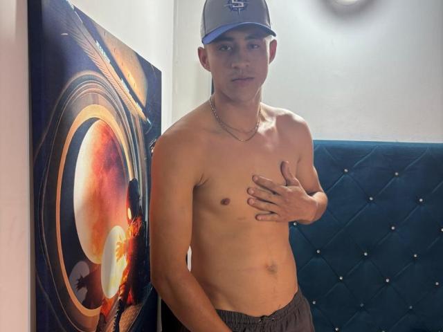 Crissteban, shirtless, wearing a gray cap, gray pants, and a chain, covering his small, flaccid, circumcised, slightly tan, average-sized cock with his hand, in a room.