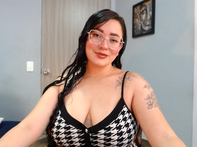 JaneAndPaull with glasses, black hair, tattoos on shoulders, wearing a houndstooth-patterned dress with black trim, showing her large tits, smiling, in a room with a door.