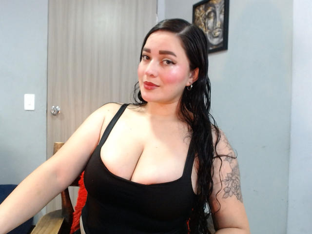 JaneAndPaull with large boobs, black tank top, glasses, and red lipstick, smiling at the camera, has tattoos on her right arm, in a room with a door.