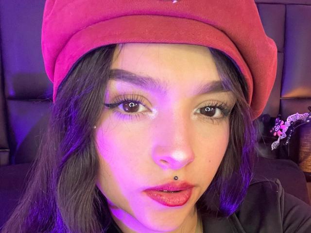 MarietteJones with long dark hair, wearing a red beret and black zip-up, holding a phone with a purple case, taking a mirror selfie, showing red lipstick.