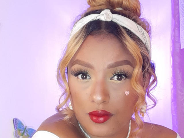 JuliaSacarf has curly blonde hair tied in a bun with a white headband. She has dark brown eyes with thick, long eyelashes and wears red lipstick. A small heart sticker is on her right cheek. She has a pearl necklace and wears a white off-the-shoulder top. The background is a soft purple, and a butterfly is partially visible on the left. The image has a warm, colorful feel.