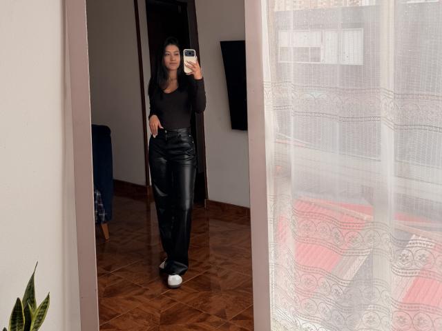 ThaliaAres with black top, black leather pants, and white sneakers, taking a mirror selfie, holding a phone, standing in a room with wooden floor, and white lace curtain.
