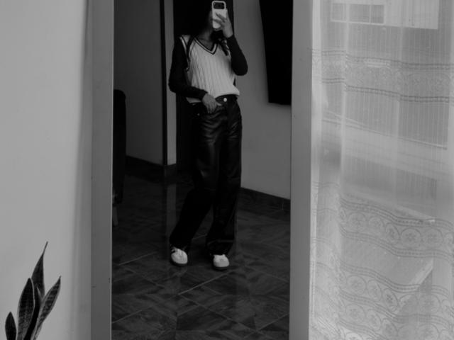 Selfie in bianco e nero di ThaliaAres, di corporatura media, che indossa una canotta bianca, pantaloni di pelle scuri e scarpe da ginnastica bianche, tiene in mano un telefono e si trova su un pavimento piastrellato..