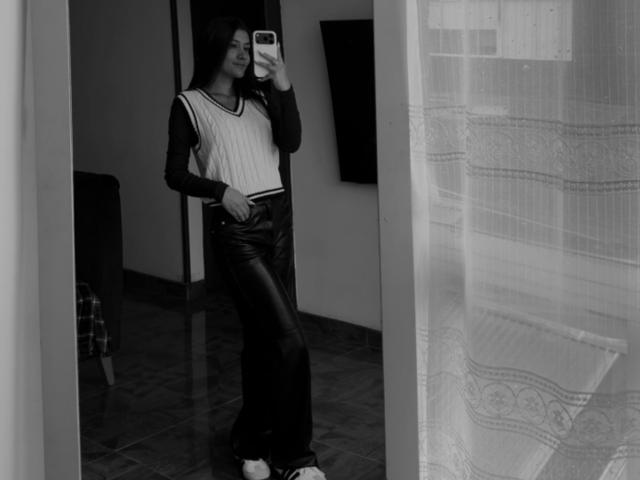Black-and-white mirror selfie of ThaliaAres with long dark hair, wearing a sleeveless white top, black leather pants, and white sneakers, standing with hand on hip.