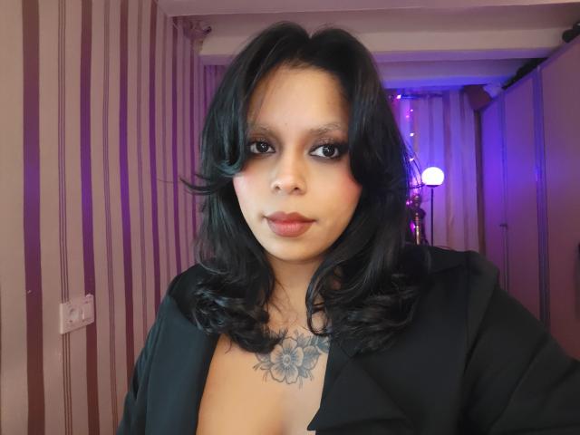 Chili, con el pelo ondulado oscuro, está en topless, vistiendo una chaqueta negra, exponiendo un tatuaje floral en el pecho, mirando directamente a la cámara. cámara.