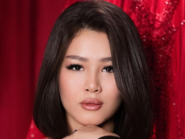 ArianaFlower has straight, dark brown hair that falls just above her shoulders. Her face is centered in the image with a slight downward gaze. She has large, dark eyes with thick, black eyeliner and long lashes. Her full, pink lips are slightly parted. She wears a black top, and her right hand is resting near her chin. The background is a rich, red textured curtain. The image has a glossy, professional look with sharp details and soft lighting.