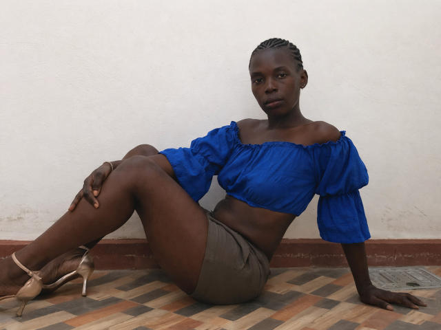 SweetApplex with dark skin, blue off-shoulder top, brown shorts, and beige heels, sitting on a tiled floor, legs crossed, arms resting, looking straight at camera.